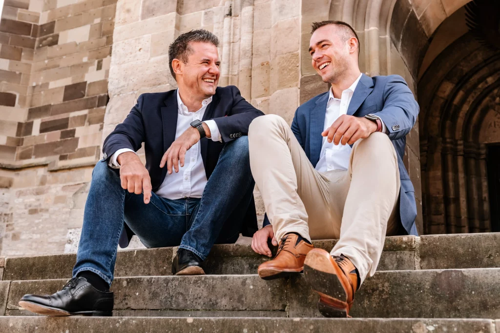 Businessportrait von 2 Männern die lachend auf einer Treppe sitzen.