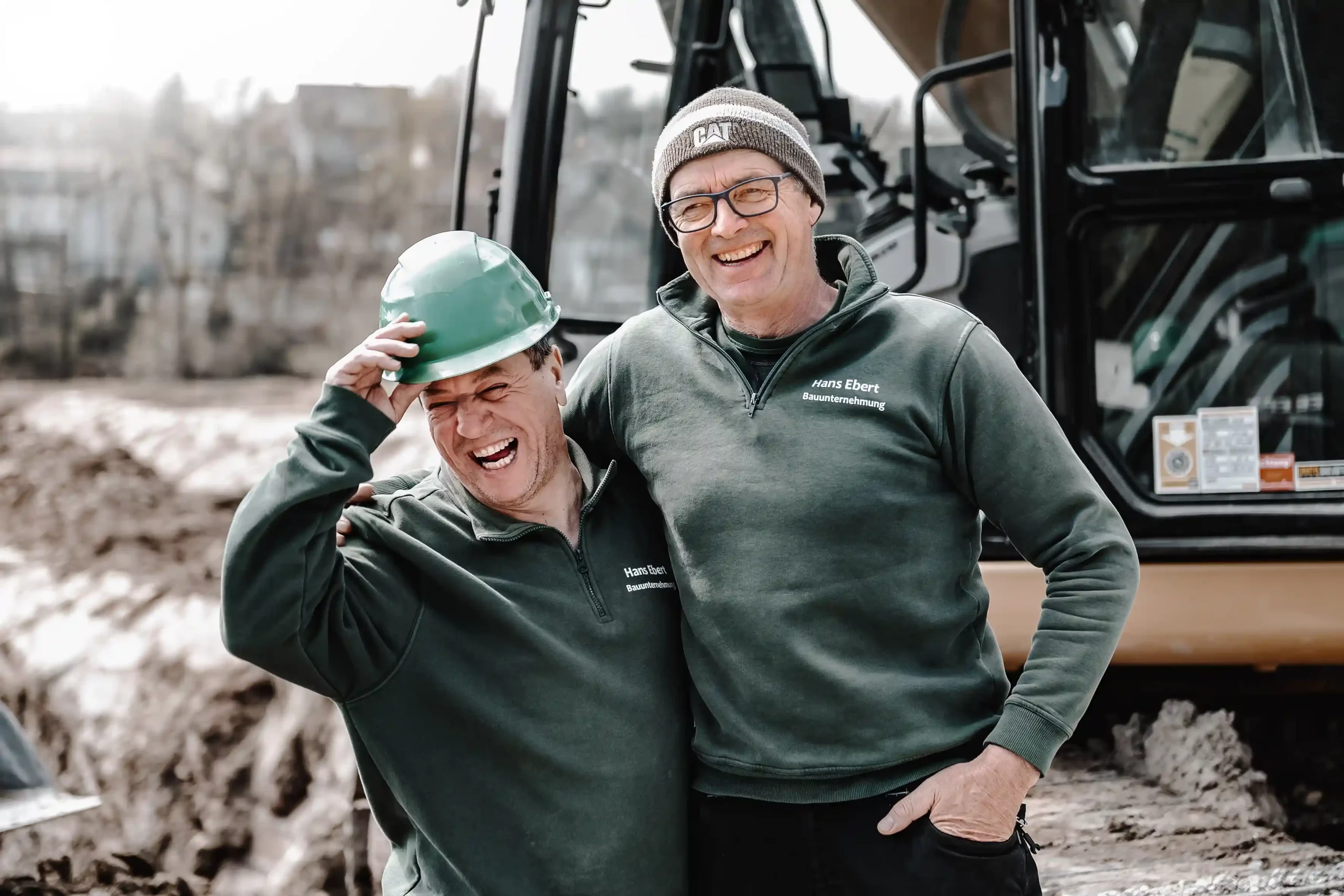 2 lachende Männer vor Bagger auf Baustelle.